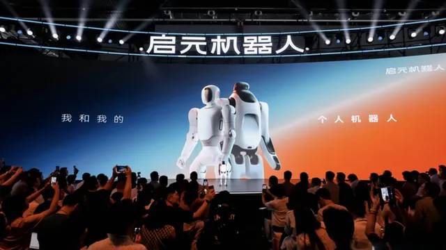 White Qiyuan Q1 and T1 robots standing side by side on a lit stage under a large "Qiyuan Robot" sign at the launch event, with a crowd filming on their phones.