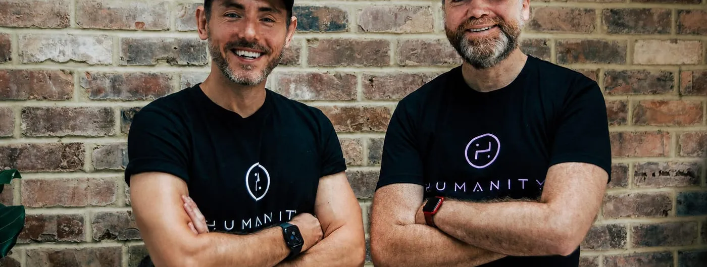 Two men stand with arms folded against a brick wall, both in black Humanity-branded caps and T-shirts, each wearing a smartwatch.