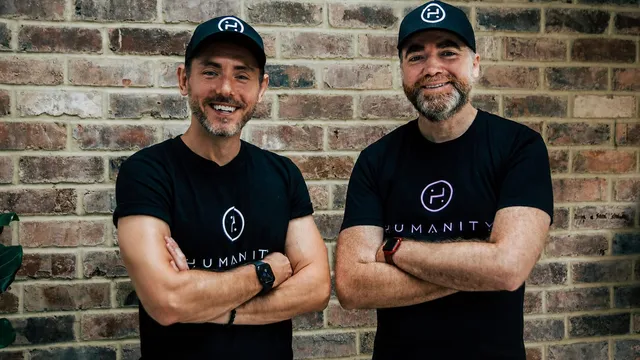 Two men stand with arms folded against a brick wall, both in black Humanity-branded caps and T-shirts, each wearing a smartwatch.