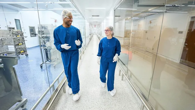 Two gowned biomanufacturing staff in blue cleanroom scrubs, hairnets, safety glasses and gloves walk and talk in a glass-walled cleanroom corridor lined with bioprocessing equipment.