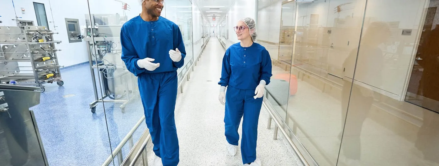 Two gowned biomanufacturing staff in blue cleanroom scrubs, hairnets, safety glasses and gloves walk and talk in a glass-walled cleanroom corridor lined with bioprocessing equipment.