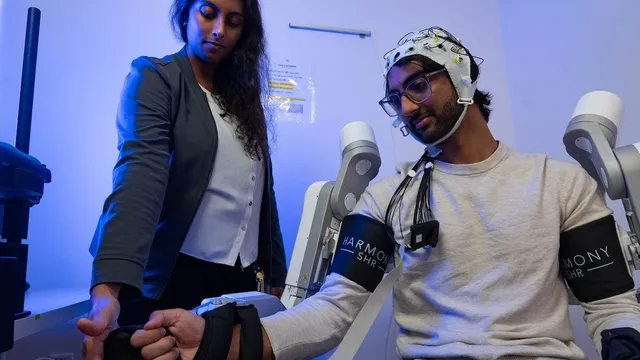 A researcher adjusts the hand strap of a seated man wearing an EEG cap and two robotic arm braces in a blue-lit lab.