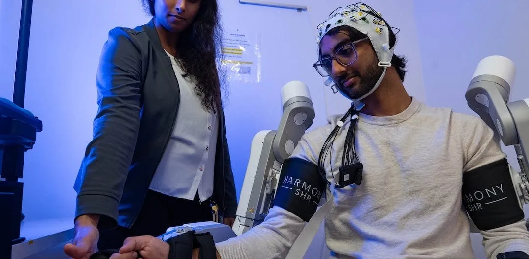 A researcher adjusts the hand strap of a seated man wearing an EEG cap and two robotic arm braces in a blue-lit lab.