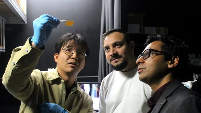 Three RMIT researchers examine the neuromorphic vision chip, a small orange square held aloft with tweezers by a gloved hand, in a darkened lab.