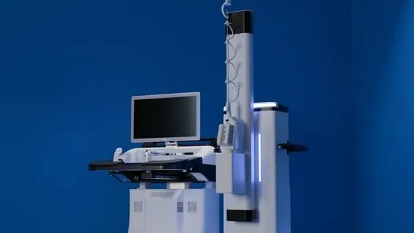 A two-cart surgical robot system photographed against a deep blue backdrop: a console with a flat monitor and keyboard tray beside a tall white robotic tower holding a thin instrument.