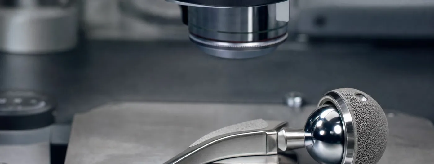 A polished metal hip replacement stem with its mirror-finish ball head and porous-coated cup lies on the stage of a laboratory microscope, the objective lens poised above it.