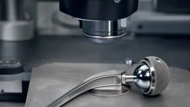 A polished metal hip replacement stem with its mirror-finish ball head and porous-coated cup lies on the stage of a laboratory microscope, the objective lens poised above it.