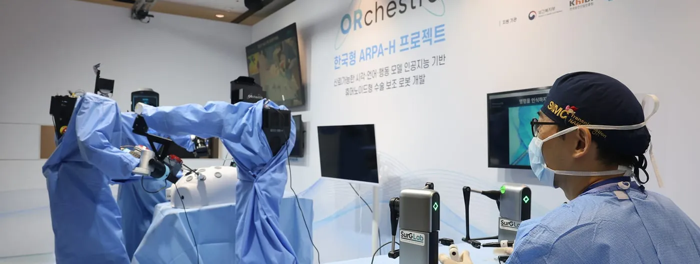 Two robotic arms draped in blue sterile covers work at an operating table while a masked surgeon watches an endoscope monitor, with the ORchestra project's partner signage on the wall behind them.