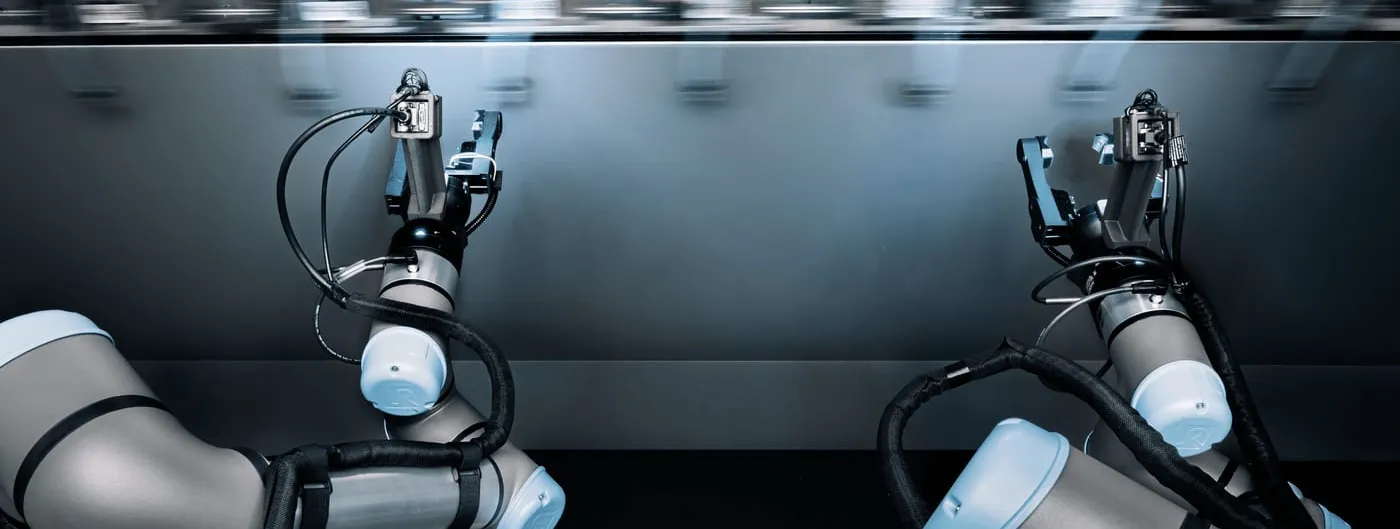 Two industrial robot arms fitted with grippers reach toward a production line that is blurred with motion behind them.