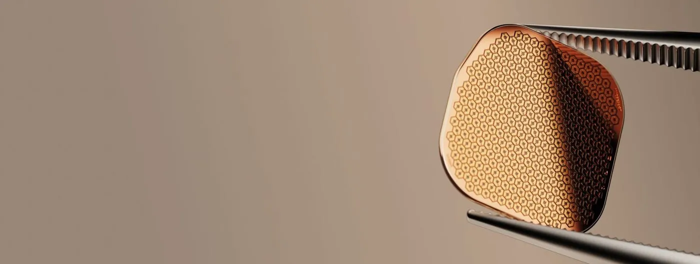 The PRIMA subretinal implant, a small rounded chip covered in a dense honeycomb of hexagonal photovoltaic pixels, held at its edge by fine surgical tweezers against a warm grey background.