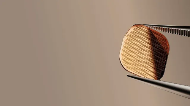 The PRIMA subretinal implant, a small rounded chip covered in a dense honeycomb of hexagonal photovoltaic pixels, held at its edge by fine surgical tweezers against a warm grey background.