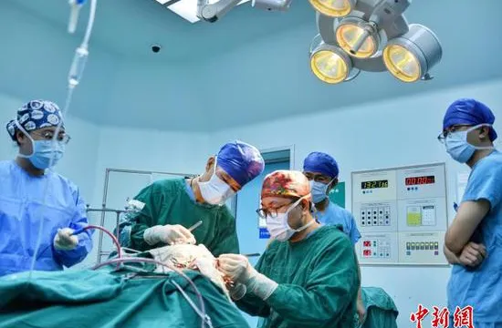 Five surgeons in caps and masks stand around a draped patient under a surgical lamp in an operating theatre, a vital-signs monitor mounted on the wall behind them.