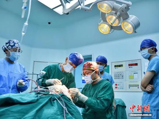 Five surgeons in caps and masks stand around a draped patient under a surgical lamp in an operating theatre, a vital-signs monitor mounted on the wall behind them.