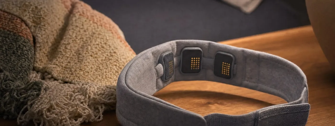 A soft grey fabric sensing headband with three slim electrode contacts on its inner face rests on a wooden bedside table beside a folded knitted throw, lit by a warm lamp.