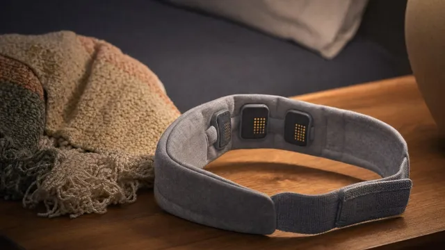A soft grey fabric sensing headband with three slim electrode contacts on its inner face rests on a wooden bedside table beside a folded knitted throw, lit by a warm lamp.