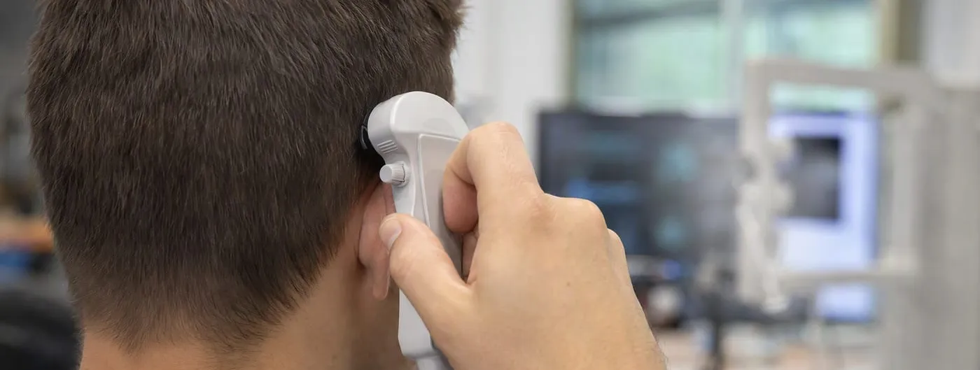 Photorealistic close-up from behind of a hand holding a white handheld ultrasound transducer device against the side of a person's head, in a research lab, with no face visible.