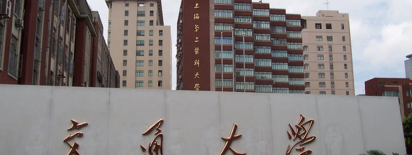 The white stone entrance wall of Shanghai Jiao Tong University, carrying the gold characters 交通大学, with the medical campus tower blocks rising behind it.
