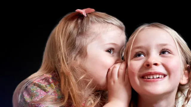 Two young girls against a dark background: one leans in with a cupped hand to whisper into the ear of the other, who smiles and looks upward.
