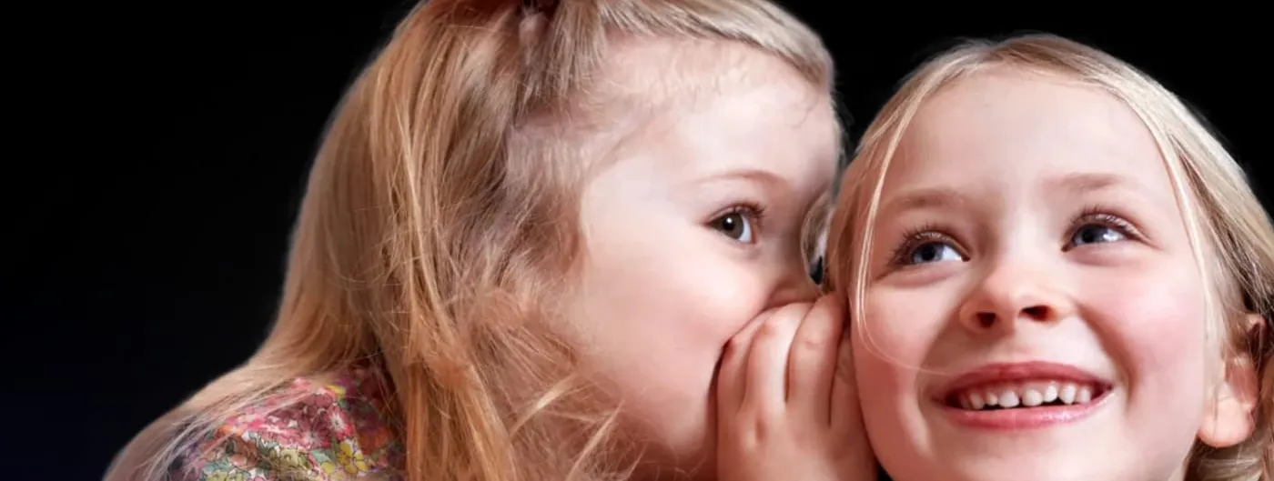 Two young girls against a dark background: one leans in with a cupped hand to whisper into the ear of the other, who smiles and looks upward.