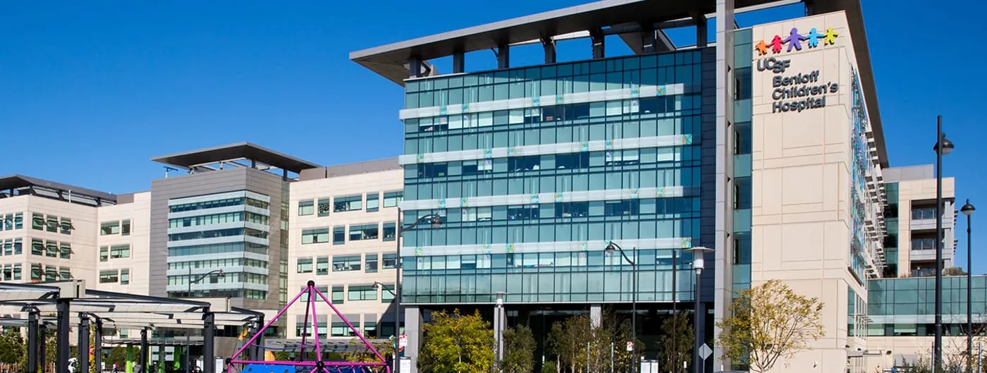 The glass-and-stone facade of UCSF Benioff Children's Hospital in San Francisco under a clear blue sky, its colourful logo on the tower and a pink-framed climbing structure in the courtyard playground.