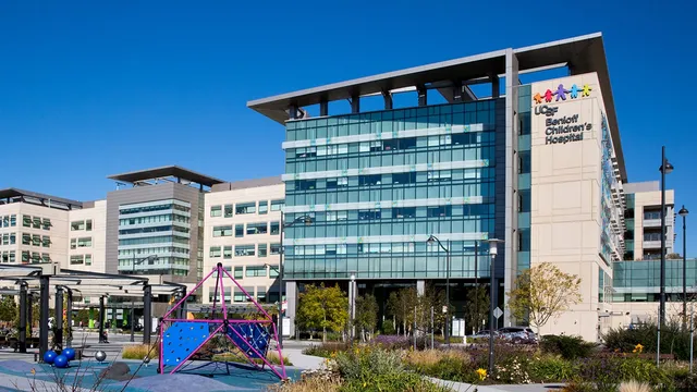 The glass-and-stone facade of UCSF Benioff Children's Hospital in San Francisco under a clear blue sky, its colourful logo on the tower and a pink-framed climbing structure in the courtyard playground.