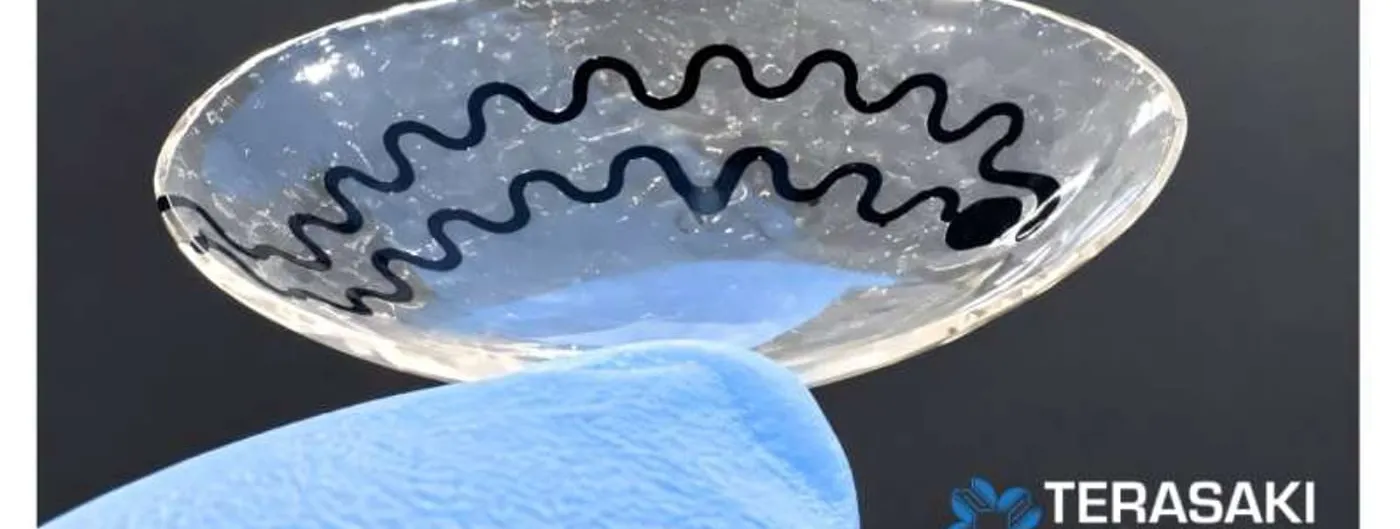 A gloved hand holds a soft, clear smart contact lens printed with a serpentine graphene-and-silver sensor trace around its rim, the Terasaki Institute logo in the corner.
