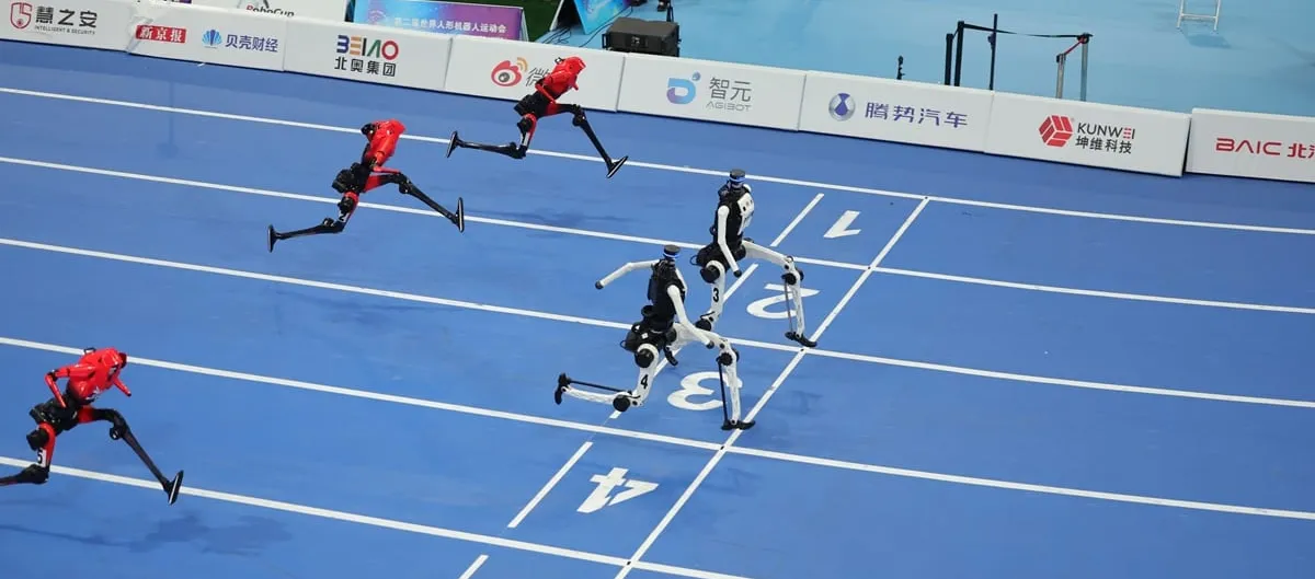 Seen from above, five humanoid robots in red and white liveries run down the marked lanes of a blue indoor track, several with both feet off the ground, sponsor boards lining the far side.