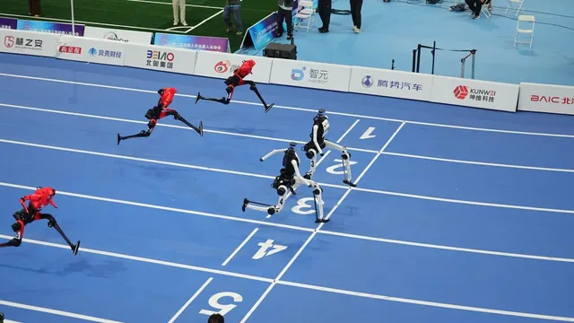 Seen from above, five humanoid robots in red and white liveries run down the marked lanes of a blue indoor track, several with both feet off the ground, sponsor boards lining the far side.