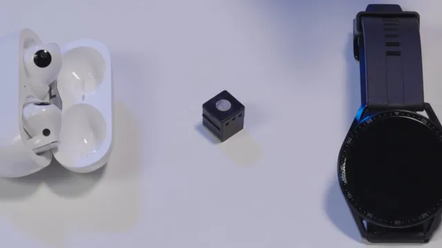 A tiny black cube-shaped EEG unit with a small round electrode pad on top, sitting on a white surface between a pair of wireless earbuds in their case and a smartwatch, to show how small it is.