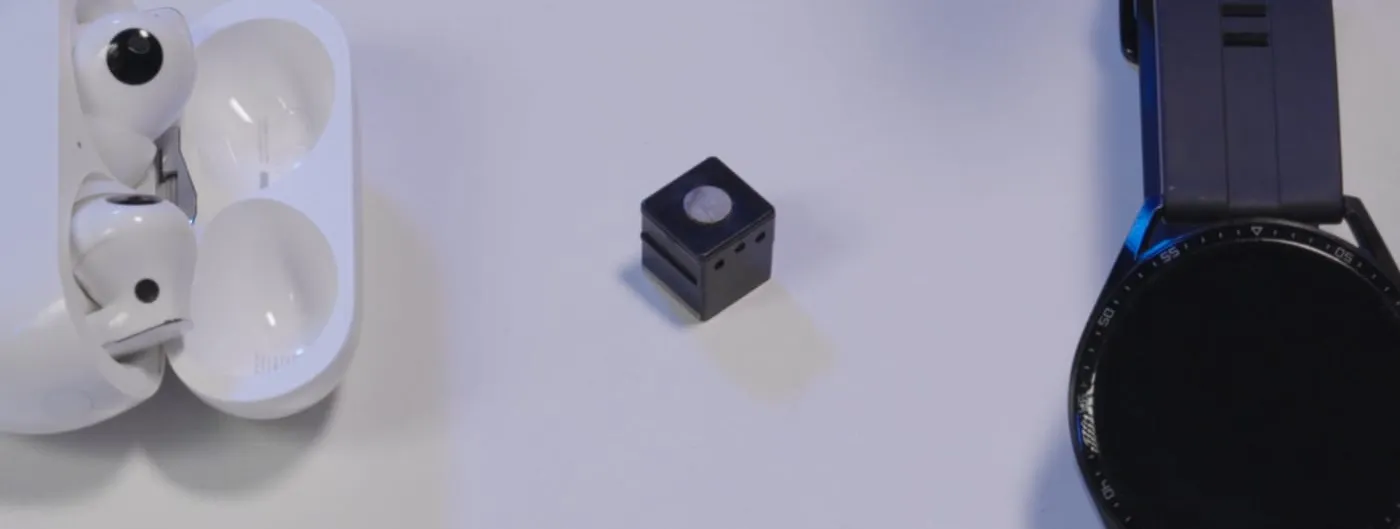 A tiny black cube-shaped EEG unit with a small round electrode pad on top, sitting on a white surface between a pair of wireless earbuds in their case and a smartwatch, to show how small it is.