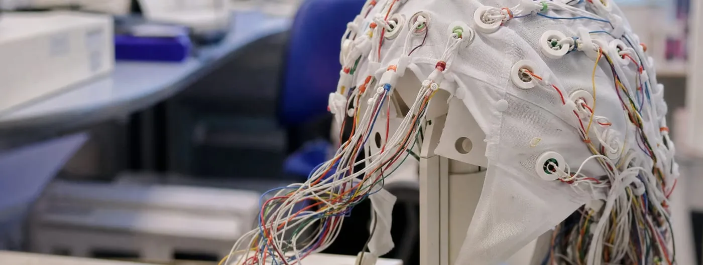 An electrode cap sits on a white stand on a clinic bench, dozens of coloured leads running from its sensors down to a connector box, with monitors and a chair out of focus behind.