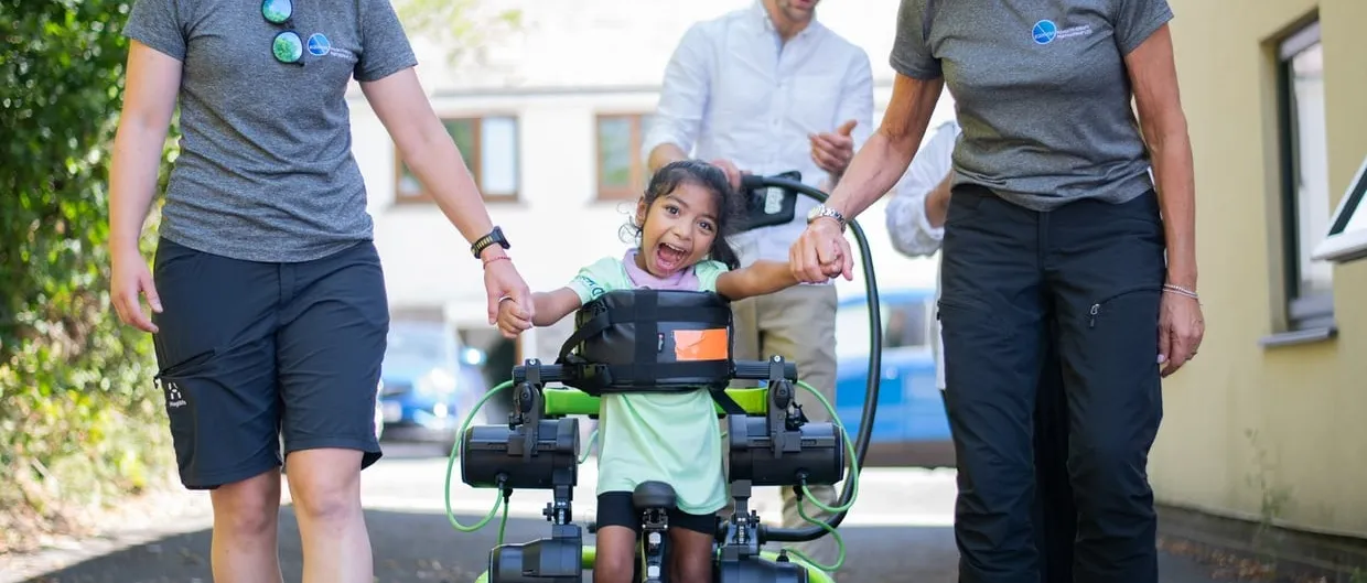 A grinning young girl walks in a green motorised leg exoskeleton mounted on a wheeled frame, holding hands with two physiotherapists on either side of her.