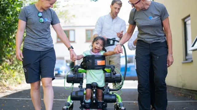 A grinning young girl walks in a green motorised leg exoskeleton mounted on a wheeled frame, holding hands with two physiotherapists on either side of her.