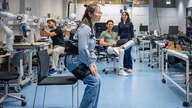 A researcher wearing a black soft hip exosuit over her jeans bends forward on the edge of a chair in a robotics lab to stand up, while three colleagues work and watch in the background.