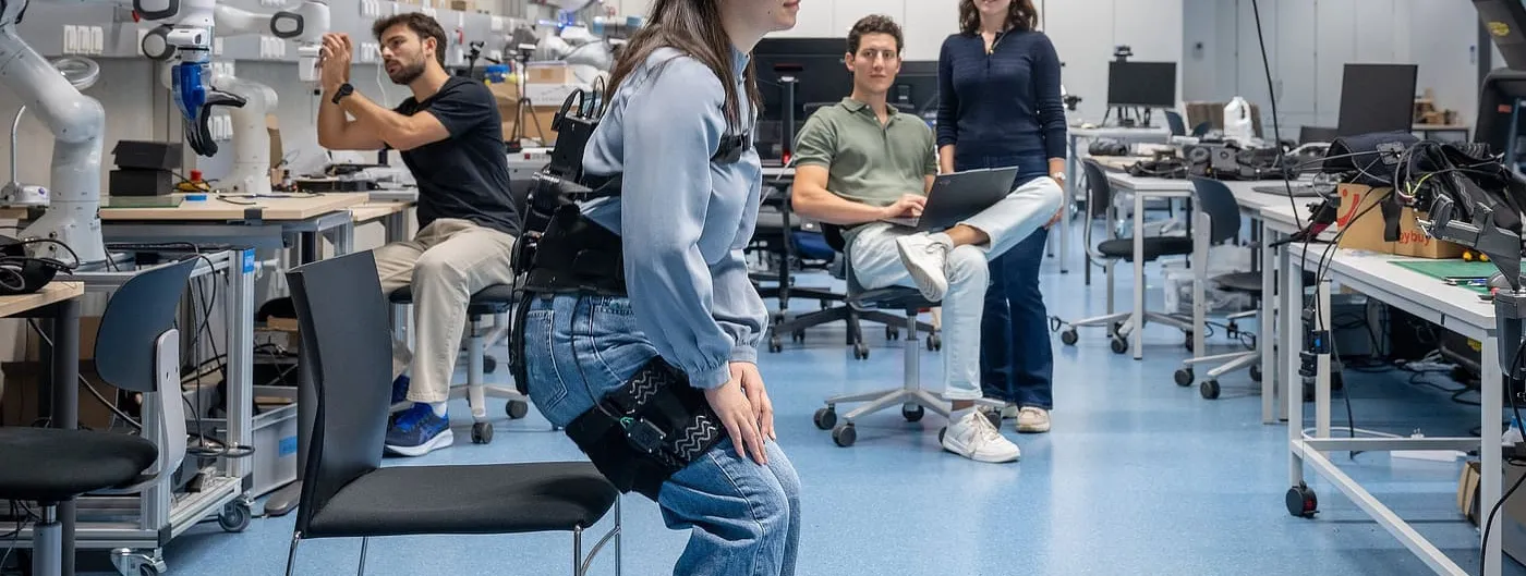 A researcher wearing a black soft hip exosuit over her jeans bends forward on the edge of a chair in a robotics lab to stand up, while three colleagues work and watch in the background.
