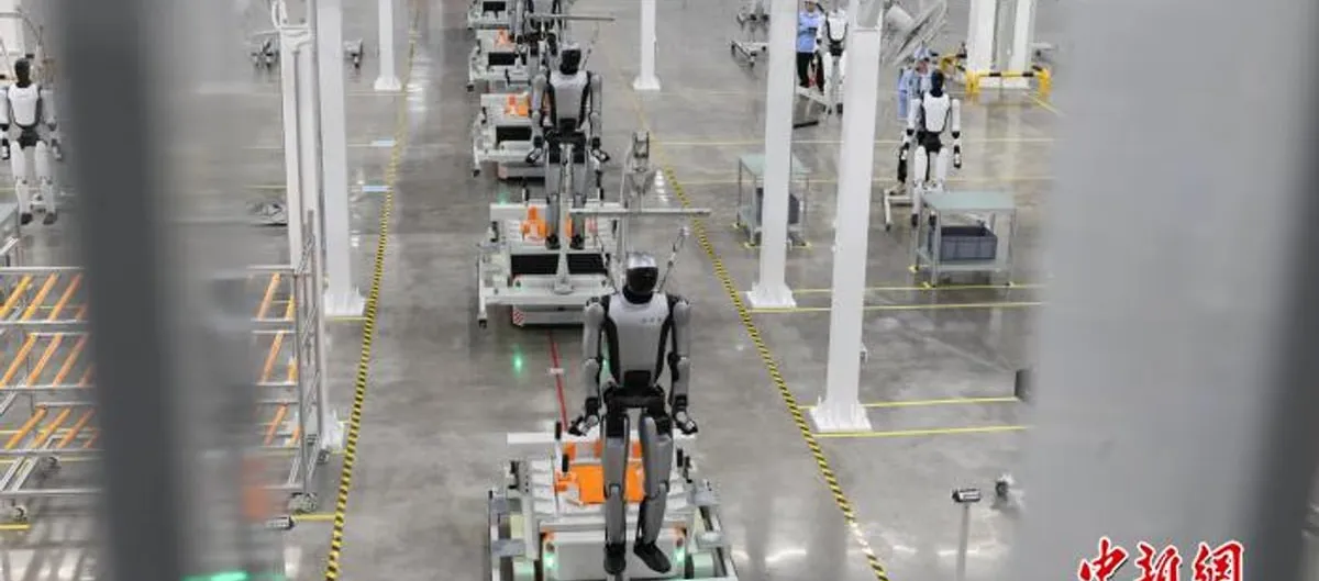 A row of white-and-black UBTECH humanoid robots stand on wheeled production carts along a factory aisle at the Liuzhou plant, with more robots and a technician visible in the background.