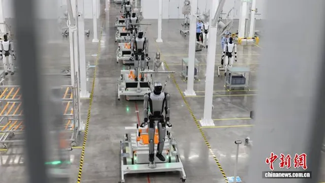 A row of white-and-black UBTECH humanoid robots stand on wheeled production carts along a factory aisle at the Liuzhou plant, with more robots and a technician visible in the background.
