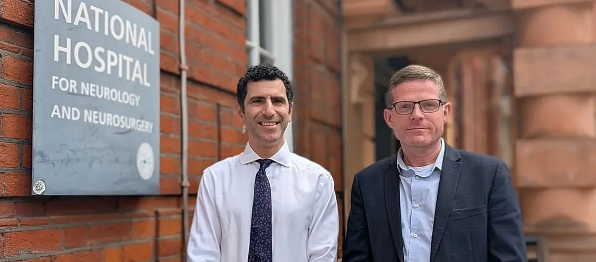 Two men in shirts and jackets stand smiling outside a red brick building, beside a slate sign reading National Hospital for Neurology and Neurosurgery.