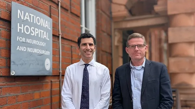 Two men in shirts and jackets stand smiling outside a red brick building, beside a slate sign reading National Hospital for Neurology and Neurosurgery.