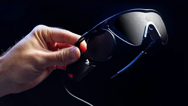 A hand holds a pair of dark wraparound glasses fitted with a camera and a trailing cable — the headset worn by recipients of a visual cortical prosthesis.