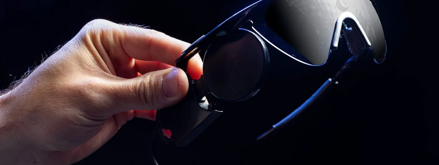 A hand holds a pair of dark wraparound glasses fitted with a camera and a trailing cable — the headset worn by recipients of a visual cortical prosthesis.
