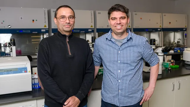 Bioengineering professors Pablo Perez-Pinera and Thomas Gaj stand side by side in their laboratory in front of a row of biosafety cabinets, with a benchtop centrifuge and racks of sample tubes beside them.