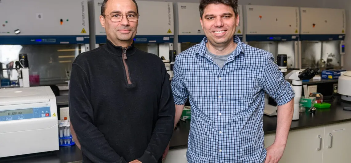 Bioengineering professors Pablo Perez-Pinera and Thomas Gaj stand side by side in their laboratory in front of a row of biosafety cabinets, with a benchtop centrifuge and racks of sample tubes beside them.