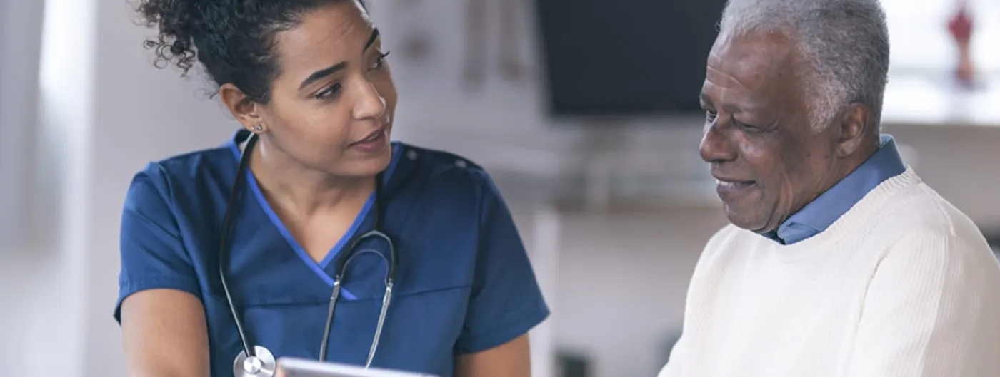 A clinician stands beside a patient, showing them information on a tablet screen in a healthcare setting.