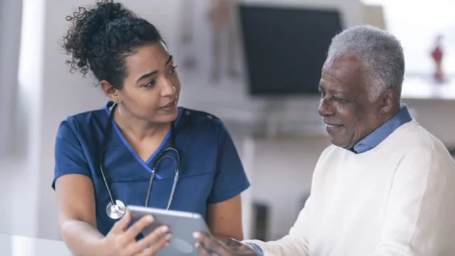 A clinician stands beside a patient, showing them information on a tablet screen in a healthcare setting.
