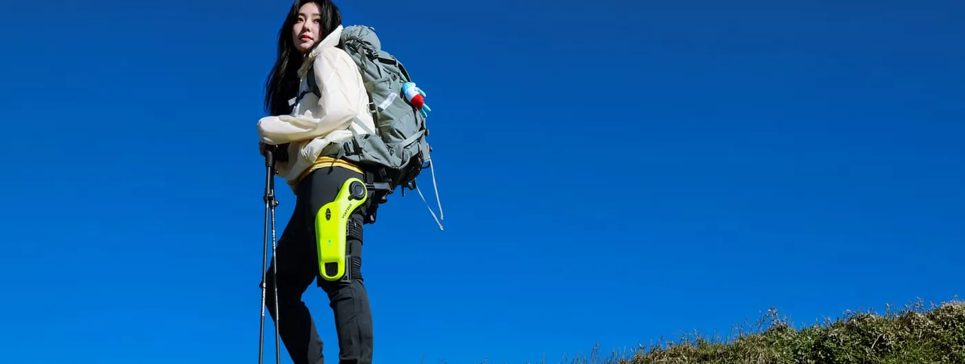 A hiker with a backpack and trekking poles stands on a grassy ridge against a deep blue sky, wearing a lime-green powered exoskeleton unit strapped to the outer thigh.