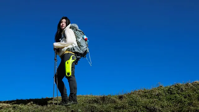 A hiker with a backpack and trekking poles stands on a grassy ridge against a deep blue sky, wearing a lime-green powered exoskeleton unit strapped to the outer thigh.