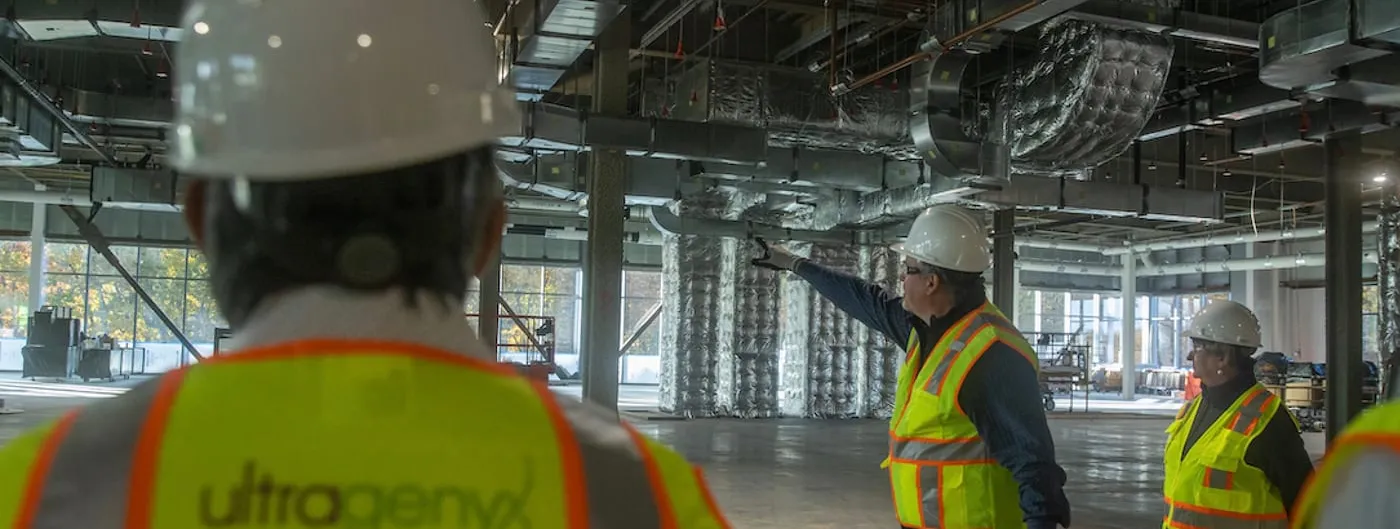 Ultragenyx employees in hard hats and high-visibility safety vests, one marked with the Ultragenyx logo, touring the open steel-and-duct interior of the company's gene therapy manufacturing facility under construction.