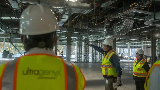 Ultragenyx employees in hard hats and high-visibility safety vests, one marked with the Ultragenyx logo, touring the open steel-and-duct interior of the company's gene therapy manufacturing facility under construction.