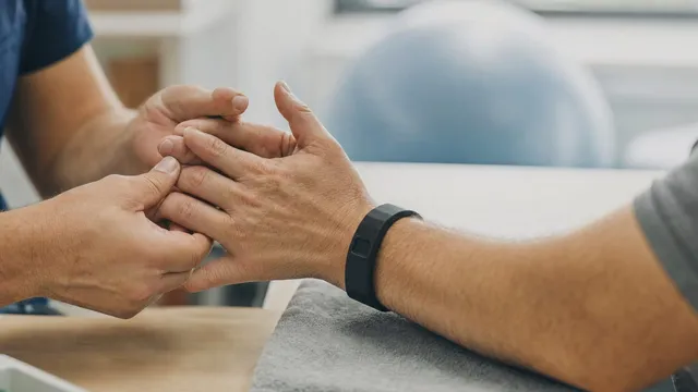 Close-up of a therapist hands guiding a patient fingers during a hand therapy session, the patient wearing a slim black wristband sensor, in a bright rehabilitation clinic with a blue exercise ball behind.